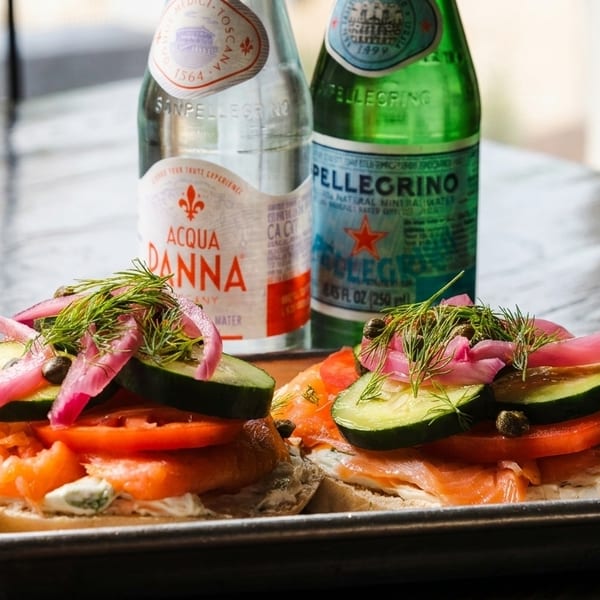 Two bagels with smoked salmon, cream cheese, cucumber, tomato, pickled onions, and dill, with two sparkling water bottles.