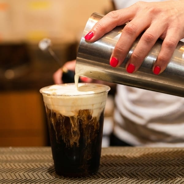 A barista pours creamy foam from a shaker into a dark beverage in a clear plastic cup.