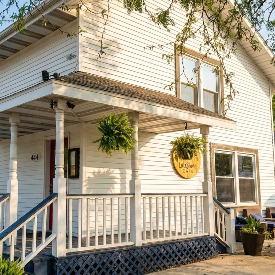 White two-story house with a porch, hanging plants, and a "LakeShore Cafe" sign, with outdoor seating.