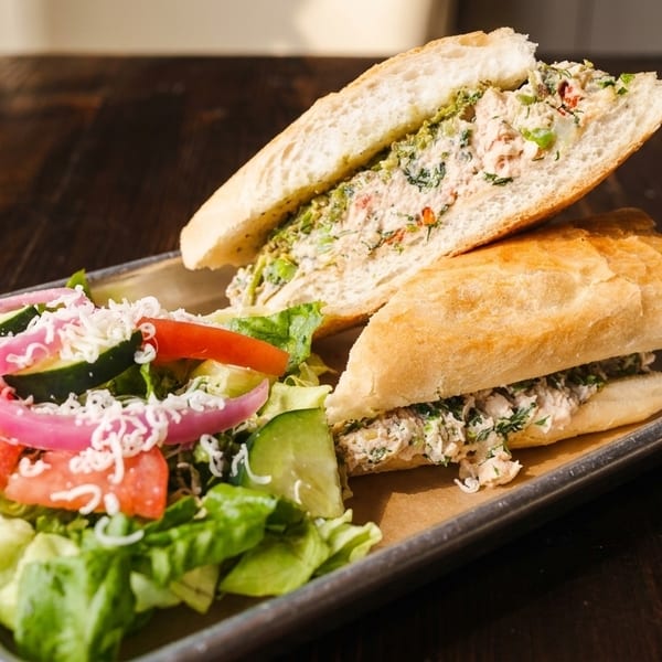 Tuna salad sandwich on a baguette with a side salad of lettuce, tomatoes, cucumbers, and red onions.
