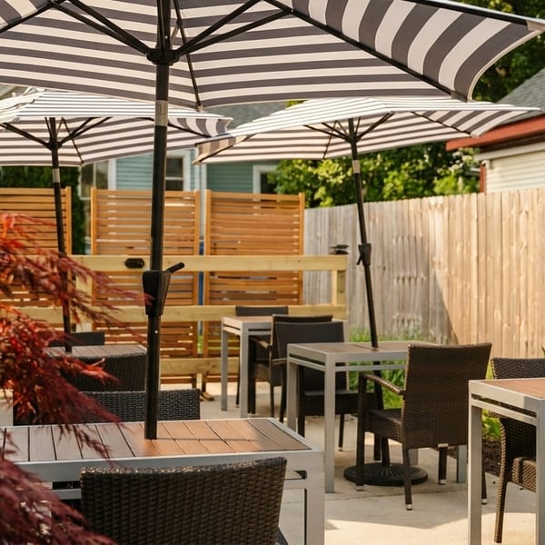 Outdoor patio dining area with striped umbrellas, square tables, and wicker chairs, surrounded by fences and foliage.