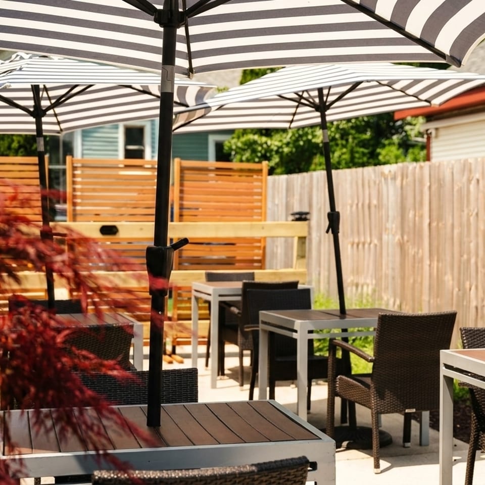 Outdoor patio dining area with striped umbrellas, modern tables, wicker chairs, and a wooden fence.